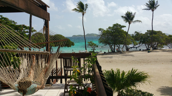 Person relaxing in a rope hammock on a wooden deck overlooking a tropical sandy beach with palm trees and turquoise ocean water.