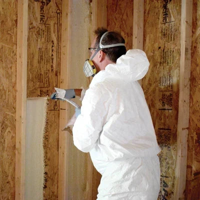 Installer wearing a white protective suit, respirator, and safety gear spraying foam insulation into wall stud cavities lined with oriented strand board (OSB).