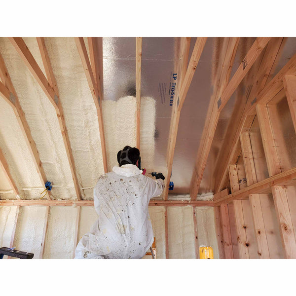 Installer wearing protective coveralls and a respirator applying spray foam insulation directly onto LP TechShield roof sheathing between wooden rafters.